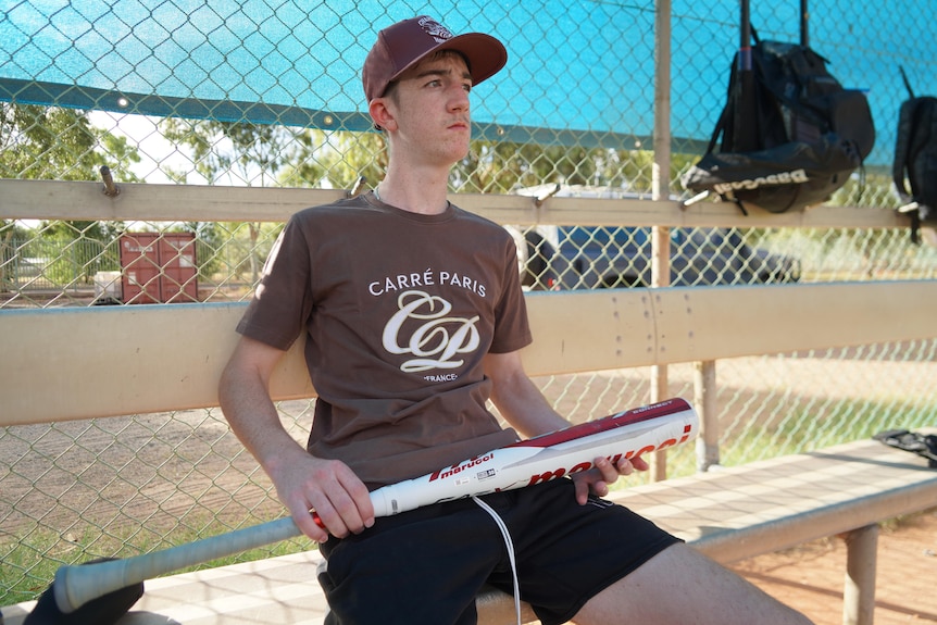 A teenage boy sits on a silver bench looking off into the distance while holding a metal baseball bat.