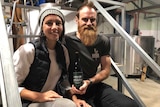 A man and a woman sitting on steel steps in a brewery, holding a bottle of beer.