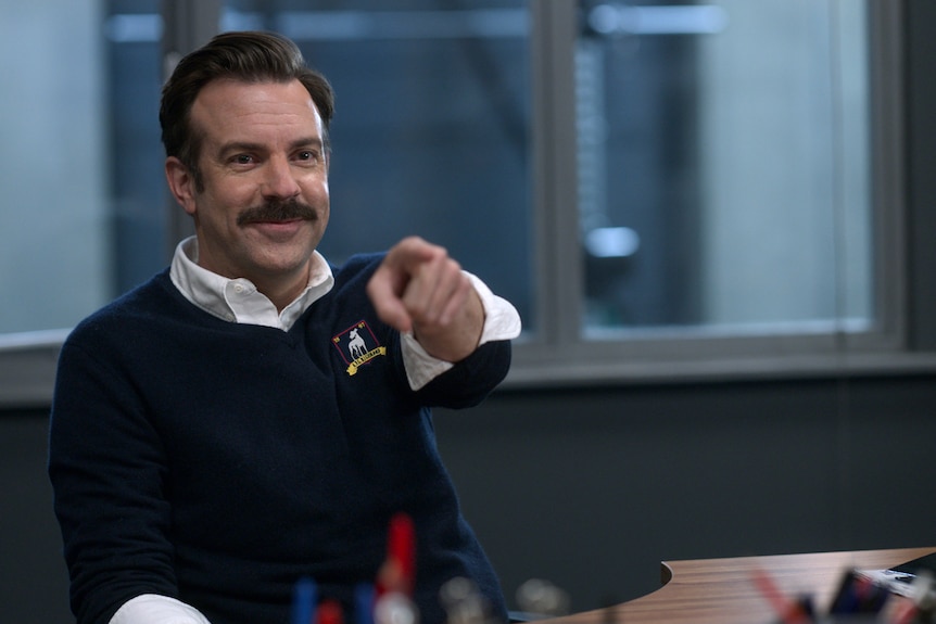 Jason Sudeikis sits at a desk and points in the Apple TV show Ted Lasso.