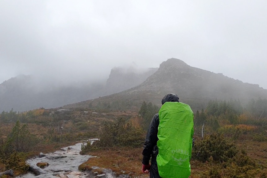 A man with a bright green hiking pack walks towards a mountain covered in thick cloud.