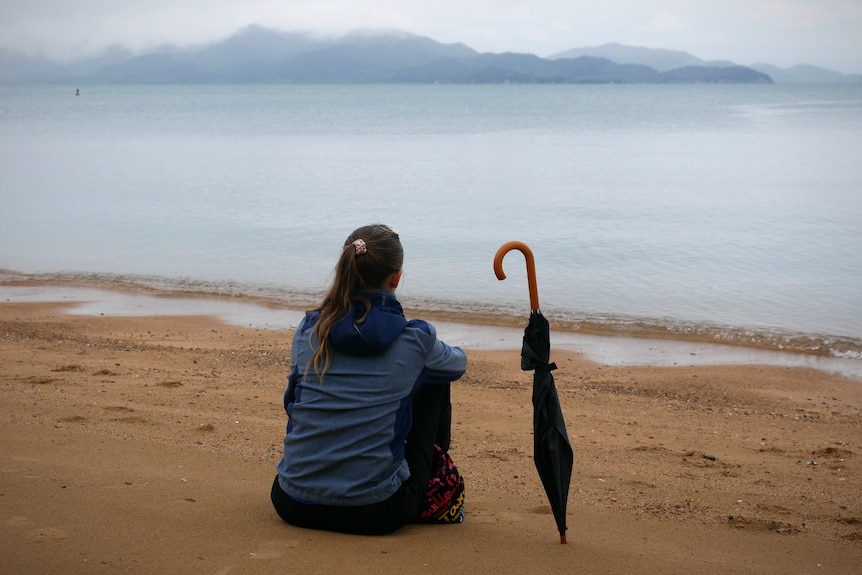 A woman sits alone on a beach with her umbrella looking out over a misty island