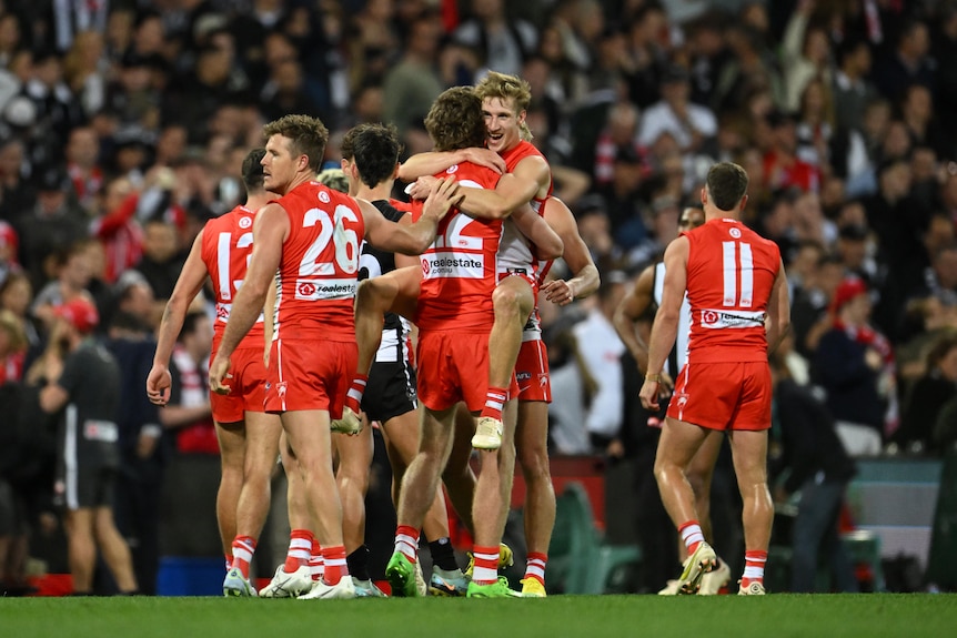 A group of Sydney Swans players hug and smile on an oval, with Collingwood players in the background.
