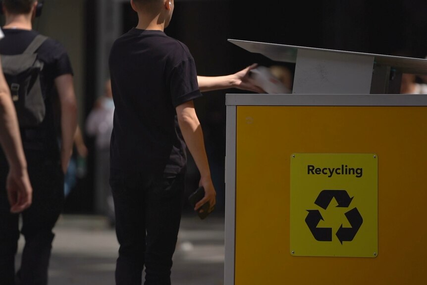 A young boy throws something into a yellow city recycling bin.
