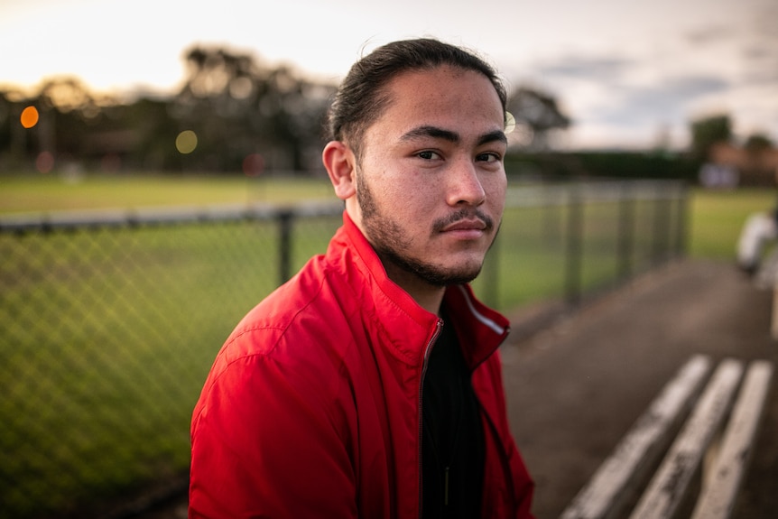 A young man wearing a red jacket sits at a bench at a sports field.