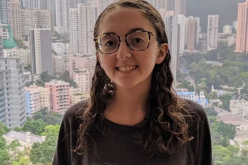 A young woman with long brown hair and glasses stands before a window overlooking Hong Kong's skyline
