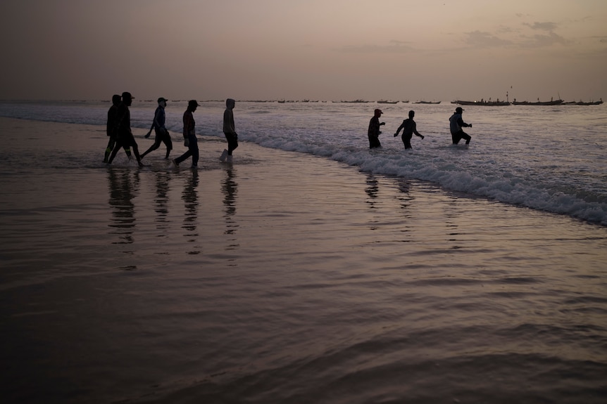 A beach is pictured at dawn or dusk as a group of young men walk into the ocean to board a fishing boat.