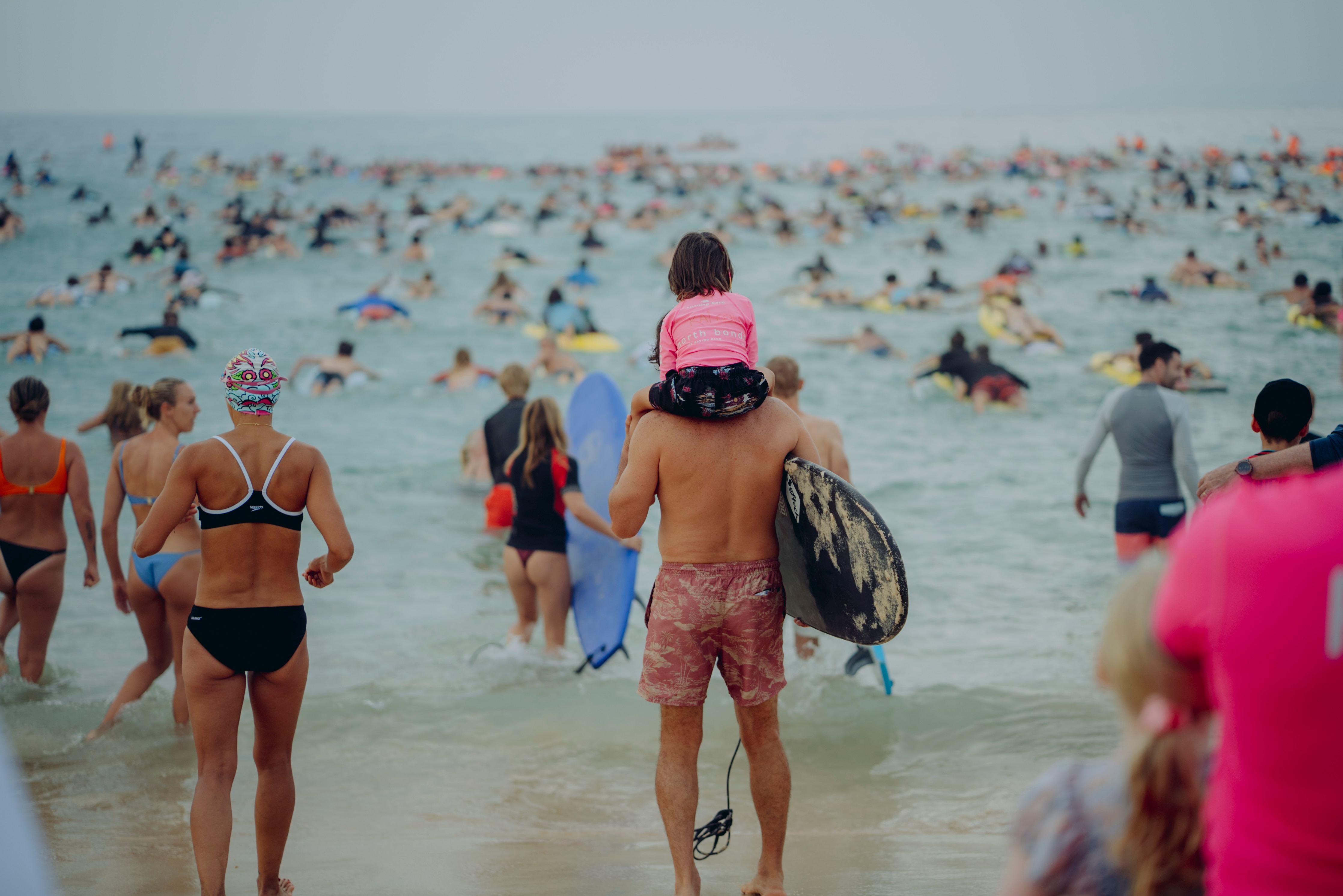 Um homem segurando uma prancha de paddle carrega seu filho no ombro enquanto se aproxima das ondas onde outros remam.
