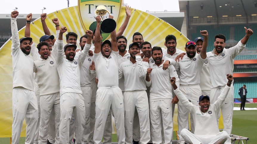Mayank Agarwal lists the trophy as his India teammates celebrate on a stage.