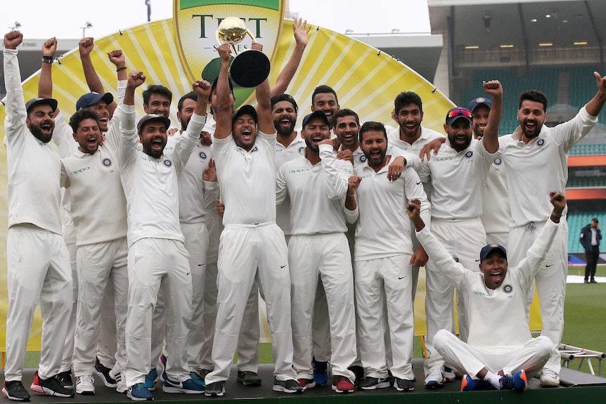 Mayank Agarwal lists the trophy as his India teammates celebrate on a stage.