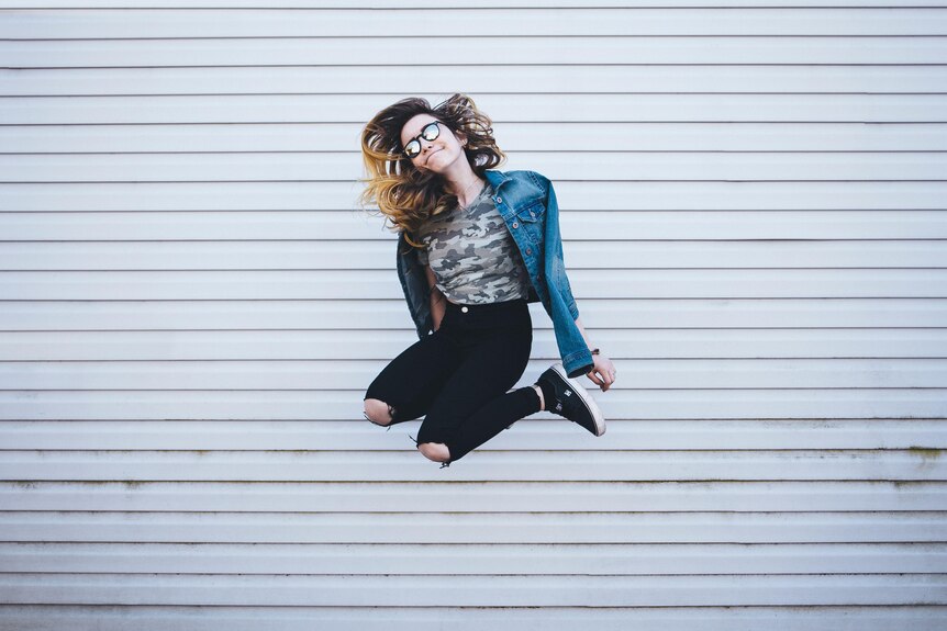 With a sheer steel background, a young woman in ripped jeans and sunglasses jumps high into the air. She's slightly smiling.