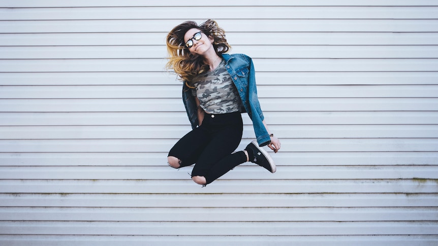 With a sheer steel background, a young woman in ripped jeans and sunglasses jumps high into the air. She's slightly smiling.