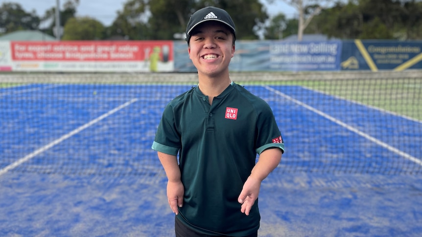 A young man with dwarfism stands on a tennis court