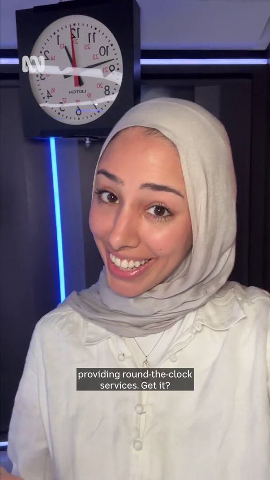 A young woman in a hijab with medium-tone skin grins, standing in front of a clock 