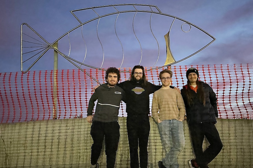 Four smiling men, one with beard, long hair, stand in front of a Sea Bass fish sculpture, plastic netting across, twilight sky.