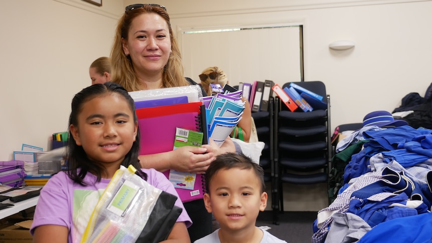 a woman smiles holding books, pencil cases, a girl and boy stand in front of her smiling