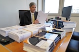 A man sitting at a desk with large documents on it and a computer