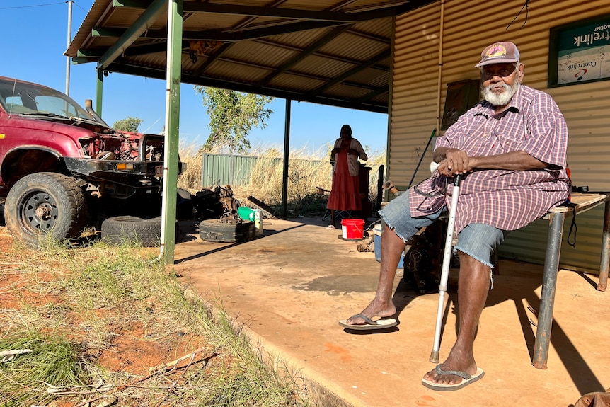 A man with a walking stick sits on a table in front of a corrugated iron house. 