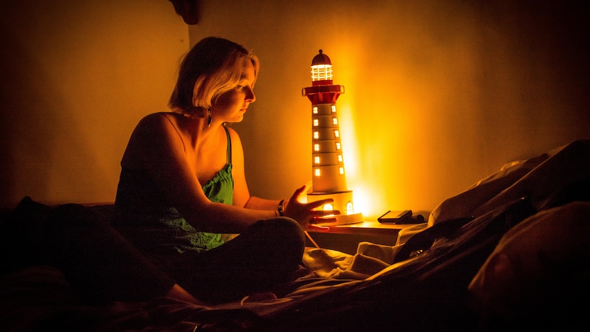 Young woman sitting on bed in the dark holding a night light, in story about managing being scared of the dark.