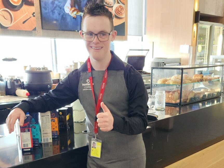 A young man in a Hudsons Coffee uniform gives a thumbs up while leaning against a cafe counter.