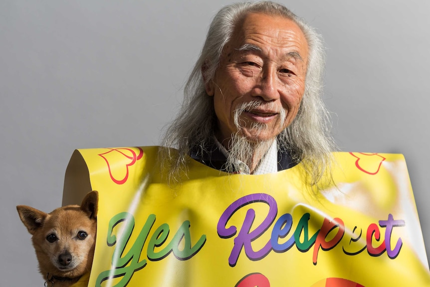 Older man stands in studio with grey hair and beard, wearing a "Yes Respect" sign, holds his dog.