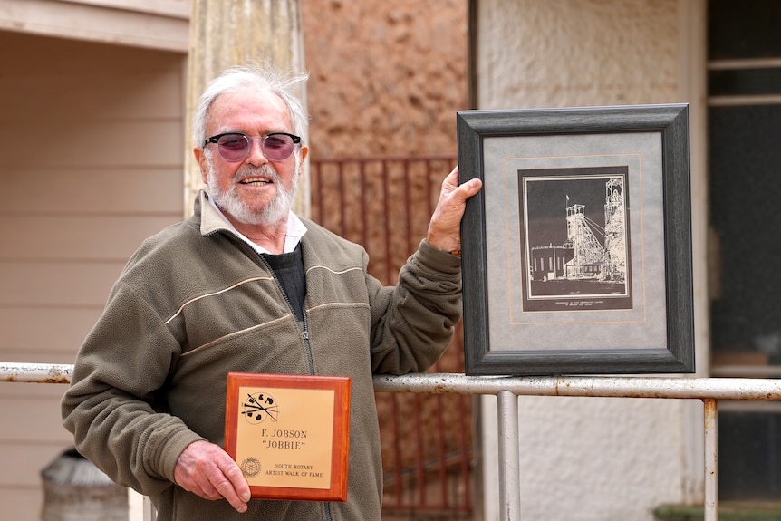 An old white man with a bear and glasses holding a painting and a plaque in Broken Hill