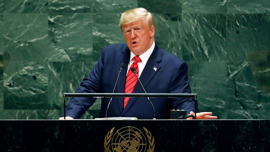 US President Donald Trump is dressed in a blue navy suit and leans over the black UN marble lectern.