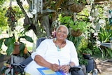 Marion Healy is sitting in a chair smiling at the camera, in front of a lush garden and plants. 