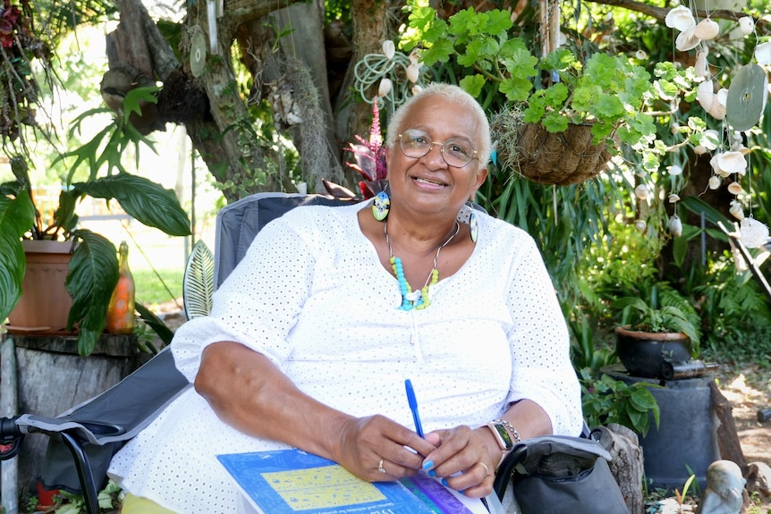 Marion Healy is sitting in a chair smiling at the camera, in front of a lush garden and plants. 