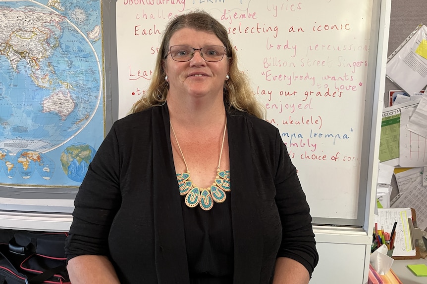 a woman with long hair and glasses smiles. there is a whiteboard behind her.