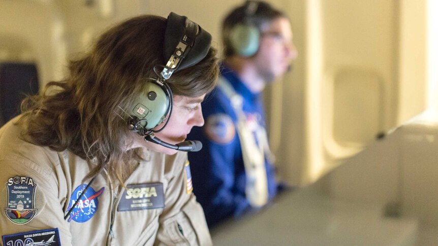 A woman wearing a NASA jumpsuit and a headset monitors a computer screen.