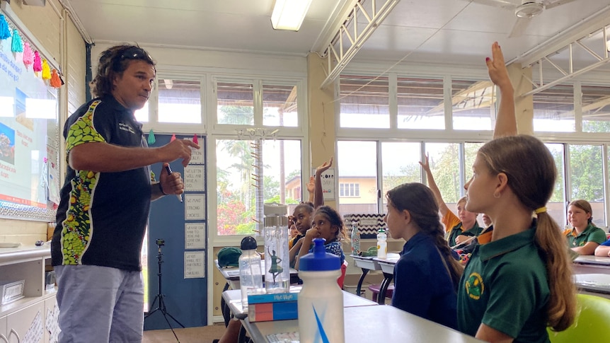 Juan Walker stands in front of a whiteboard at the front of a classroom facing students, some with hands in the air.