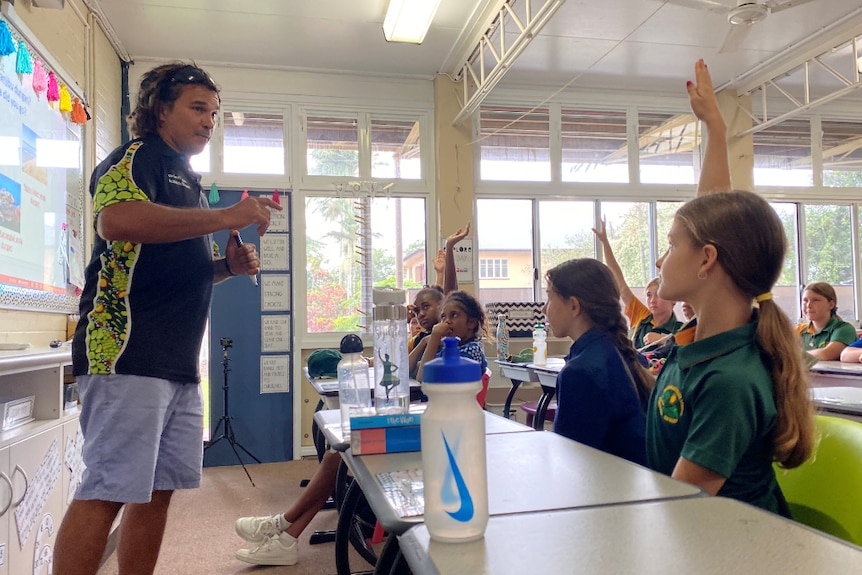 Juan Walker stands in front of a whiteboard at the front of a classroom facing students, some with hands in the air.