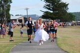 A woman in wedding dress and sneakers and a man in a suit with shorts run along a path in a park, flanked by other runners.