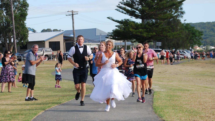 A woman in wedding dress and sneakers and a man in a suit with shorts run along a path in a park, flanked by other runners.