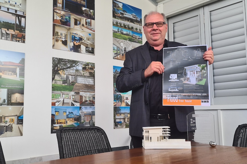 A man stands holding a sign in front of a wall of house designs in an office