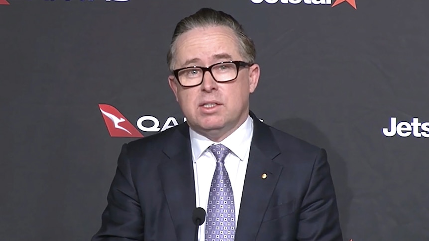 Alan Joyce sitting in front of a Qantas media backdrop while speaking into a microphone. 