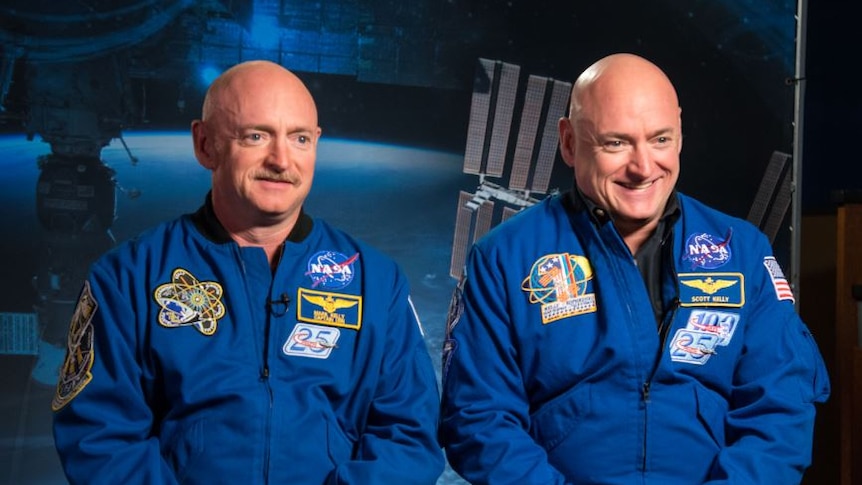 Mark and Scott Kelly sit in NASA jackets smiling in front of a blue space background.