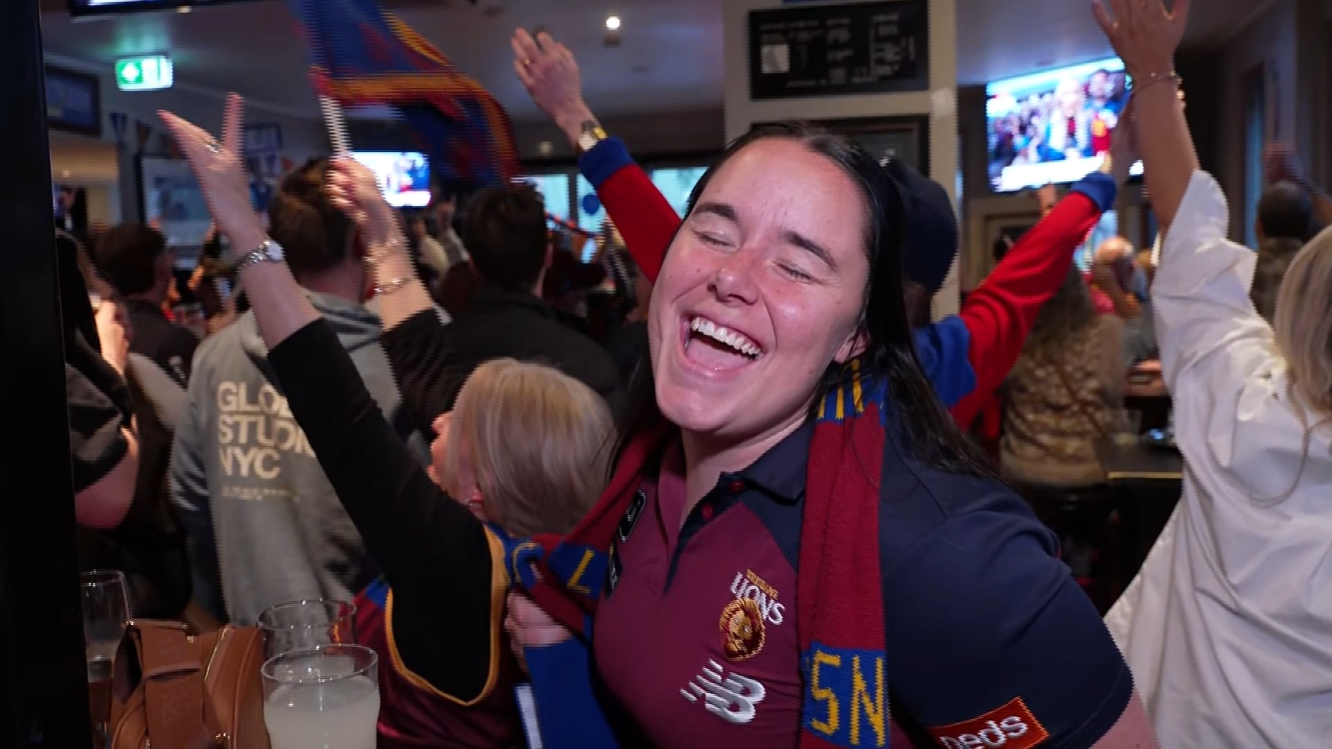 A woman with a Brisbane Lions scarf closes her eyes, singing, as people behind her hold their arms in the air.