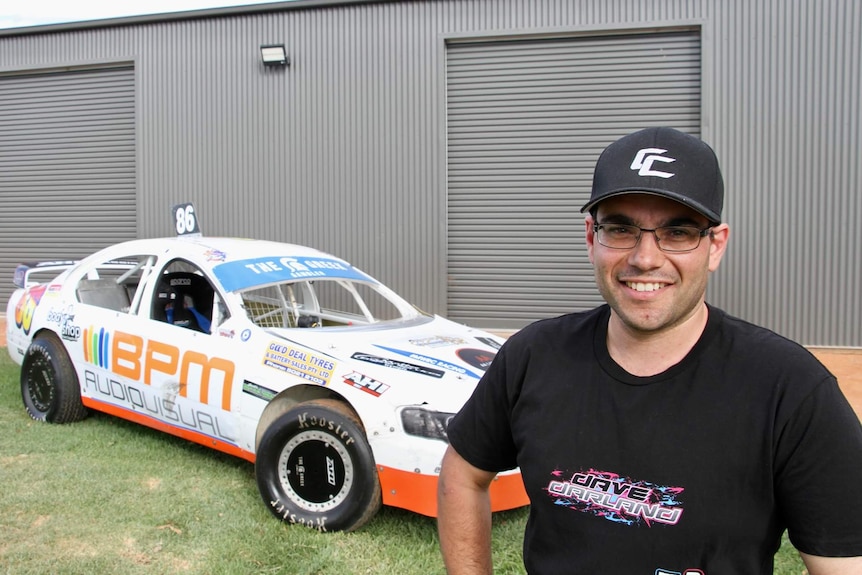 A man with glasses and a cap stands in front of a white and orange race car. 