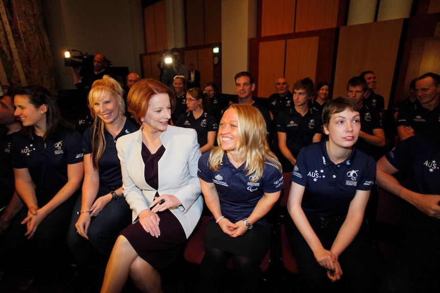 Julia Gillard in a black dress and white blazer chatting to a blonde woman in team outfit, with other players seated nearby