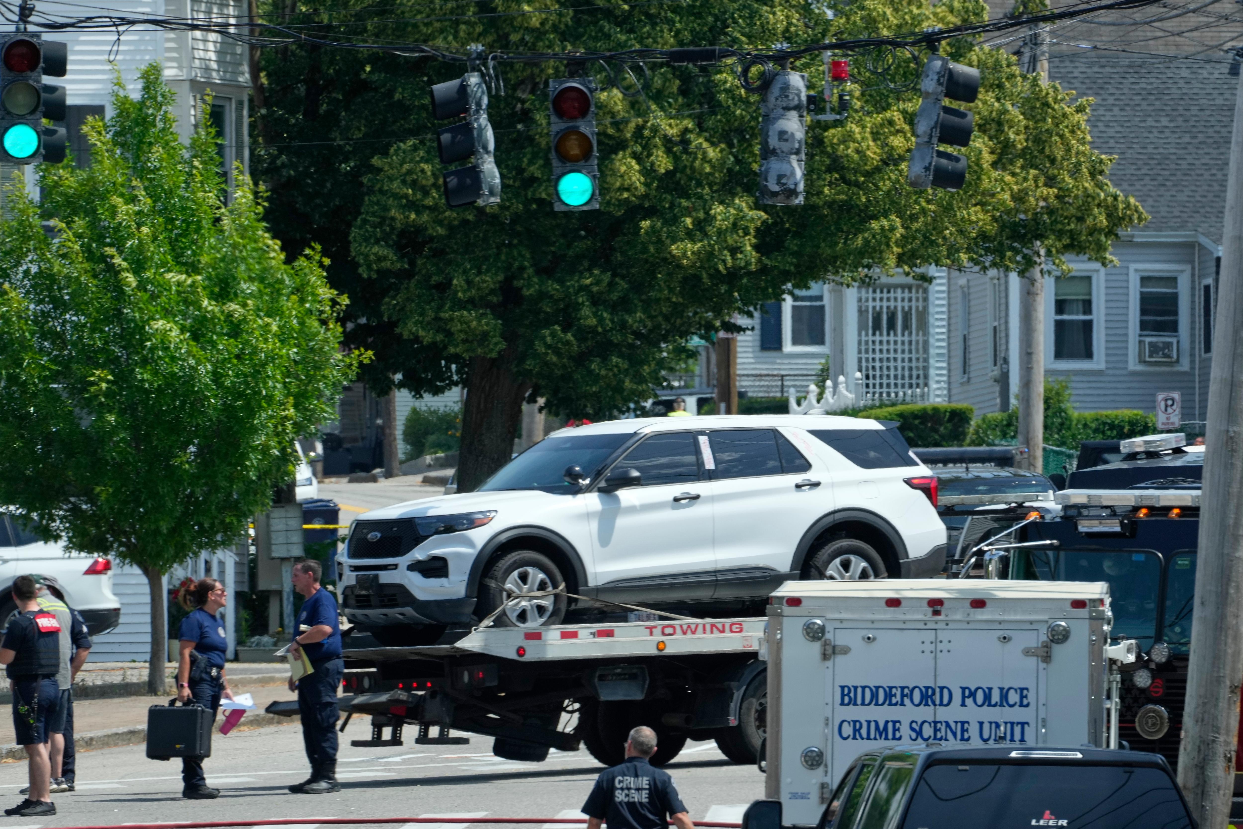 A car on a flatbed truck being towed. A trailer with the words Biddeford Police Crime Scene Unit is near.