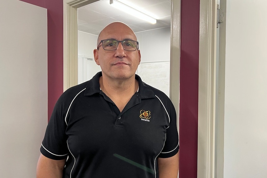 A man, wearing a black t-shirt and glasses, stands in an office.