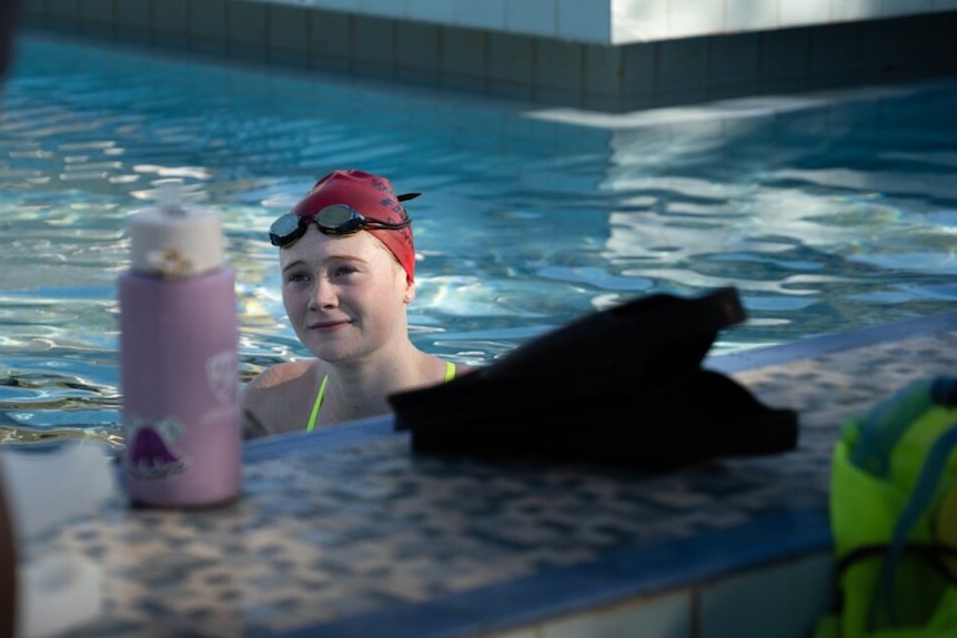 Girl wearing bright swimming cap with goggles pushed to the top of her head takes a break in the pool.