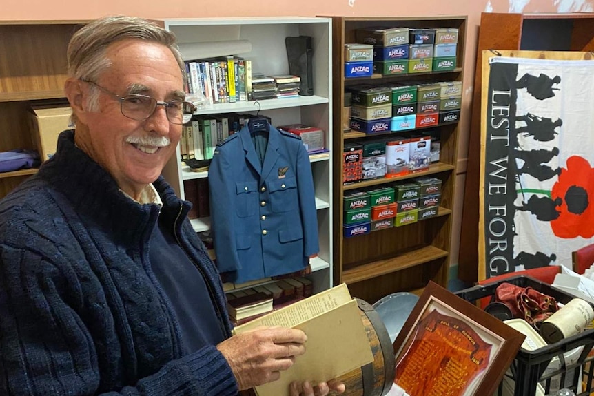 Man in foreground of messy room holding book, war memorabilia in background.