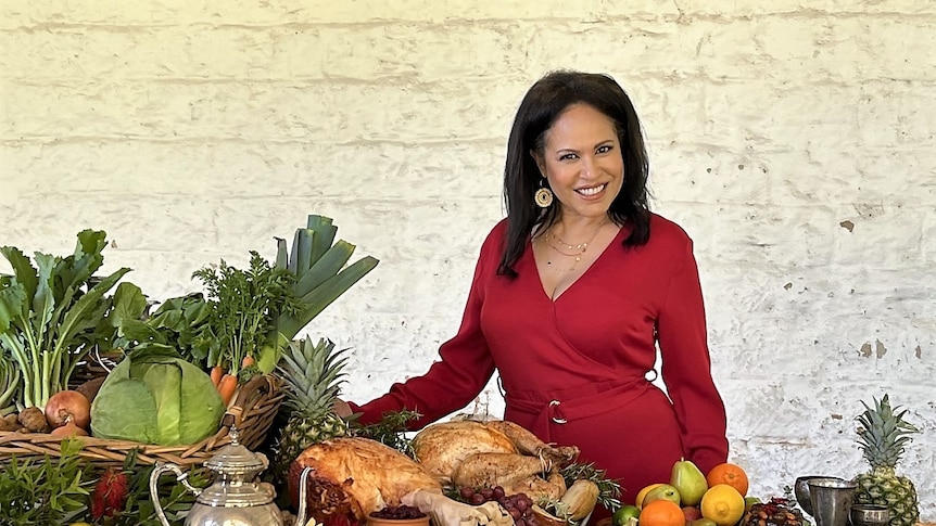 Christine Anu posing in front of a Christmas feast with Australian native flowers layed out on a table wearing a red dress.
