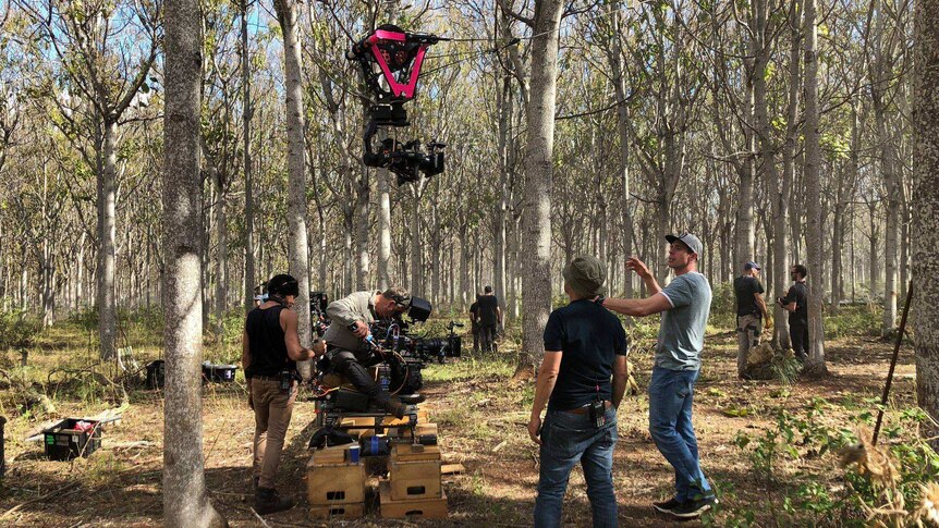People stand in a forest of trees, some are operating a movie camera on the ground and another suspended on wires in the air.