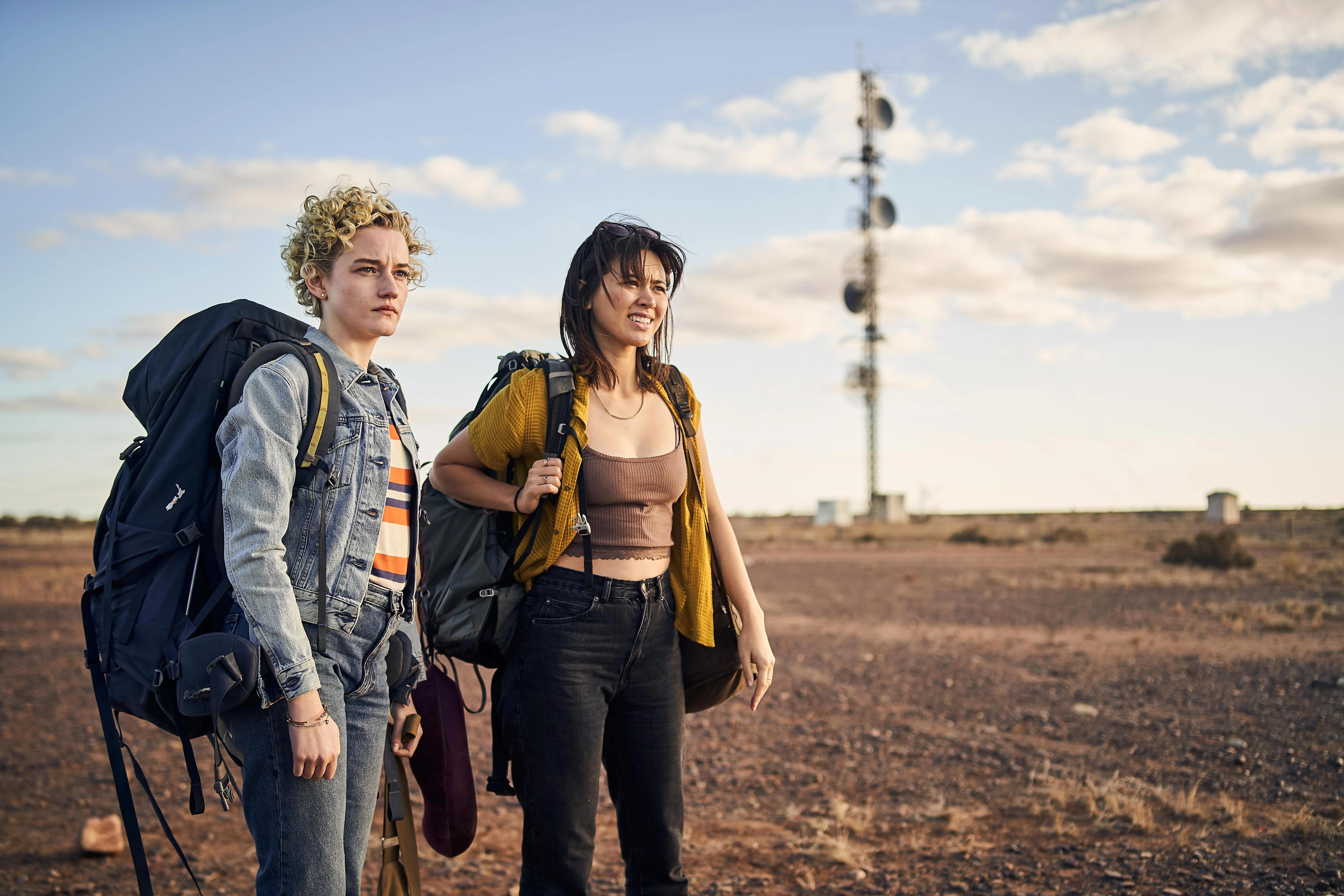 Julia Garner and Jessica Henwick squint in the outback, wearing huge backpacks, in the film The Royal Hotel.