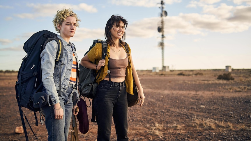 Julia Garner and Jessica Henwick squint in the outback, wearing huge backpacks, in the film The Royal Hotel.