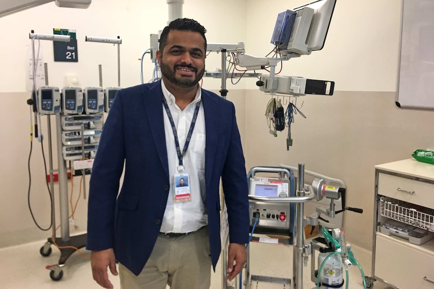 Dr Kiran Shekar stands in a hospital room surrounded by medical equipment.