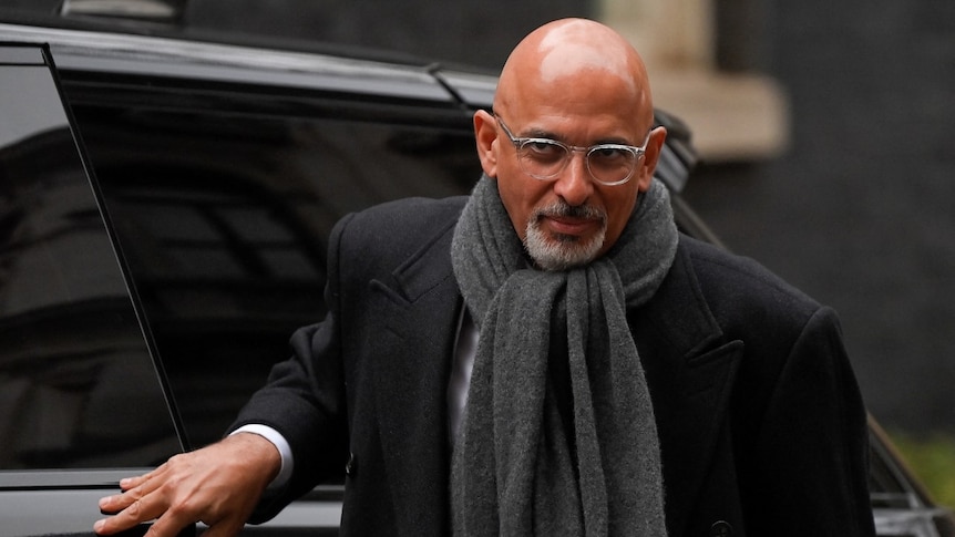 A bald man exits a black car, dressed in a black suit with a grey scarf around his neck.